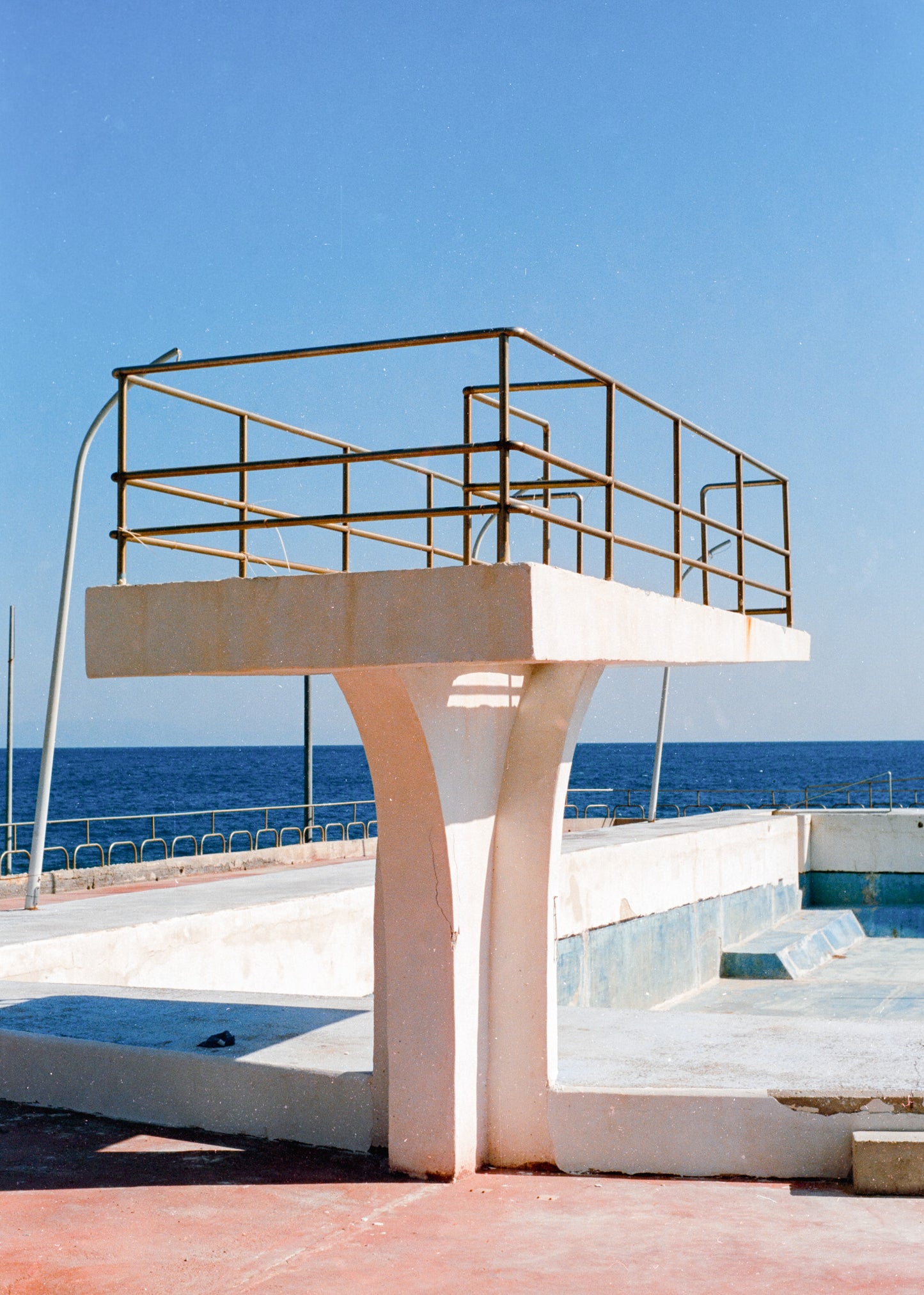 Analoges Foto als Kunstdruck. Ein Sprungturm steht vor einem leeren, verlassenen Pool. Dahinter ist das Mittelmeer zu sehen.
