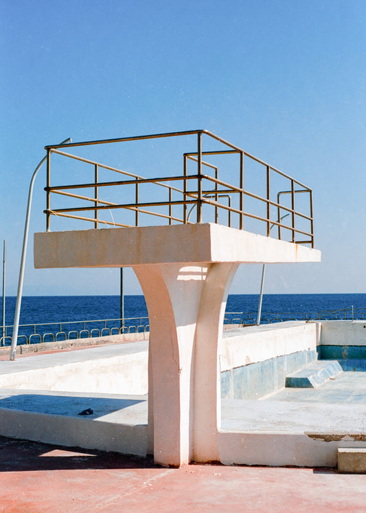 Analoges Foto als Kunstdruck. Ein Sprungturm steht vor einem leeren, verlassenen Pool. Dahinter ist das Mittelmeer zu sehen.